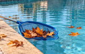 A blue pool skimmer net gathering fallen orange autumn leaves from clear swimming pool water.